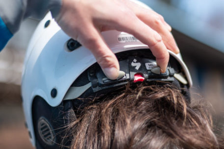 The back of a woman's head as she adjusts the width of her helmet with her left hand.