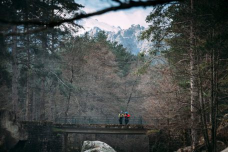 Zwei Menschen in Kajakbekleidung stehen auf einer Brücke. Im Hintergrund sind Bäume und schneebedeckte Berge.
